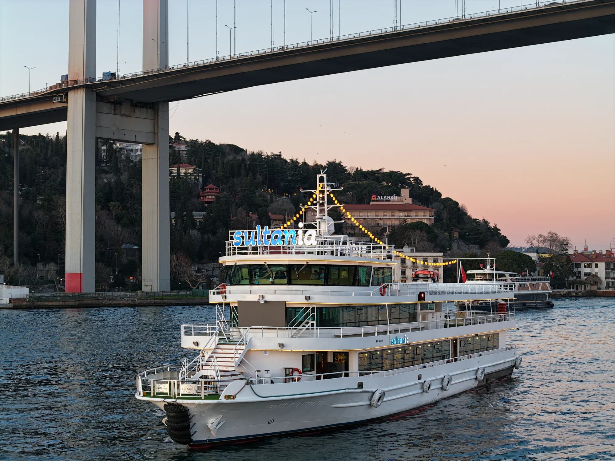 A dinner cruise boat passing under the Bosphorus Bridge at dusk