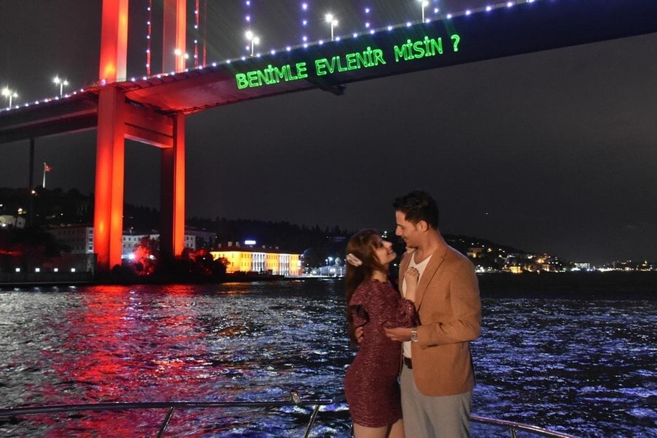 A couple embracing on the yacht's foredeck beneath the illuminated Bosphorus Bridge