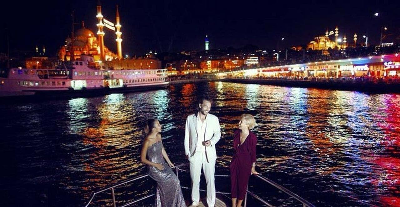 Guests on the yacht's foredeck at night with an illuminated mosque on the waterfront