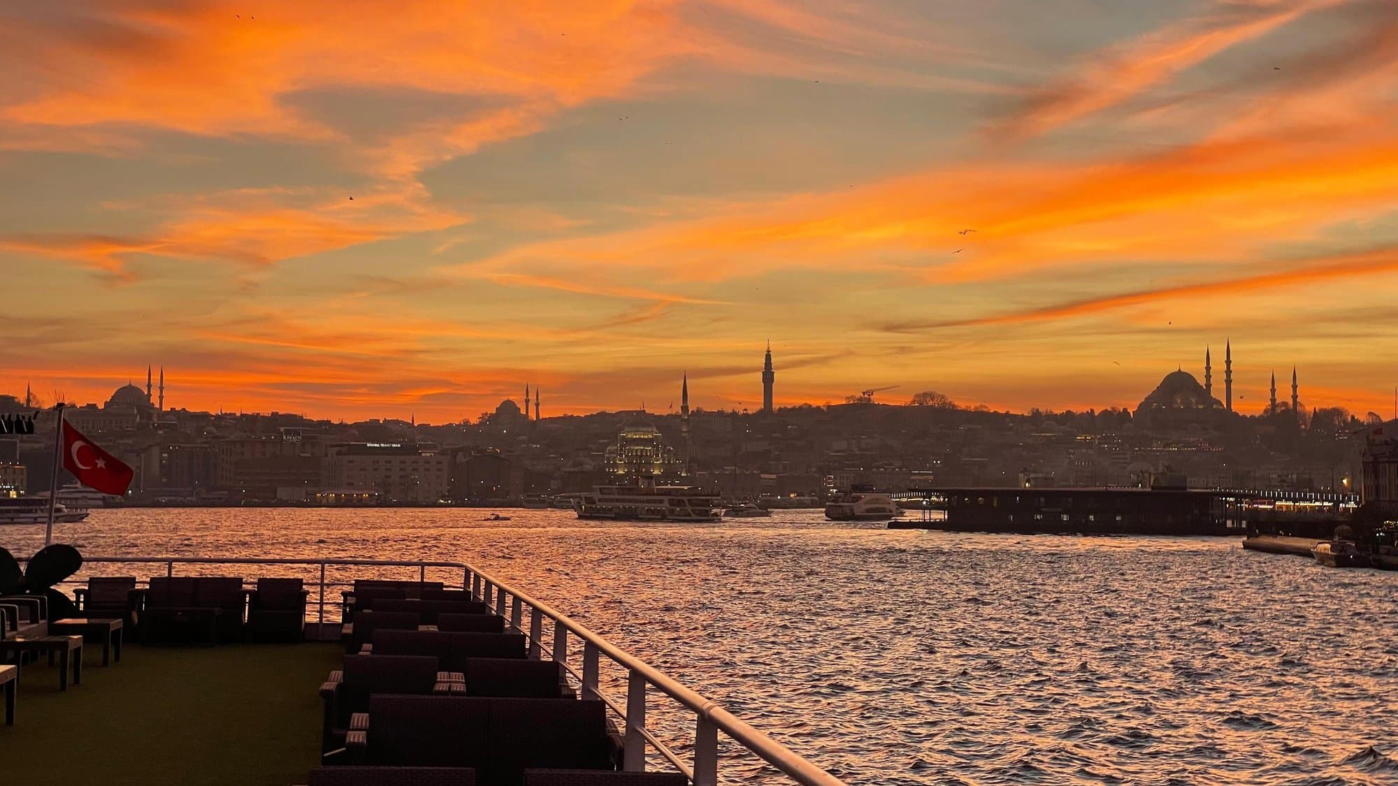 A fiery orange sunset over the historic Istanbul skyline of mosques and towers, seen from the open deck