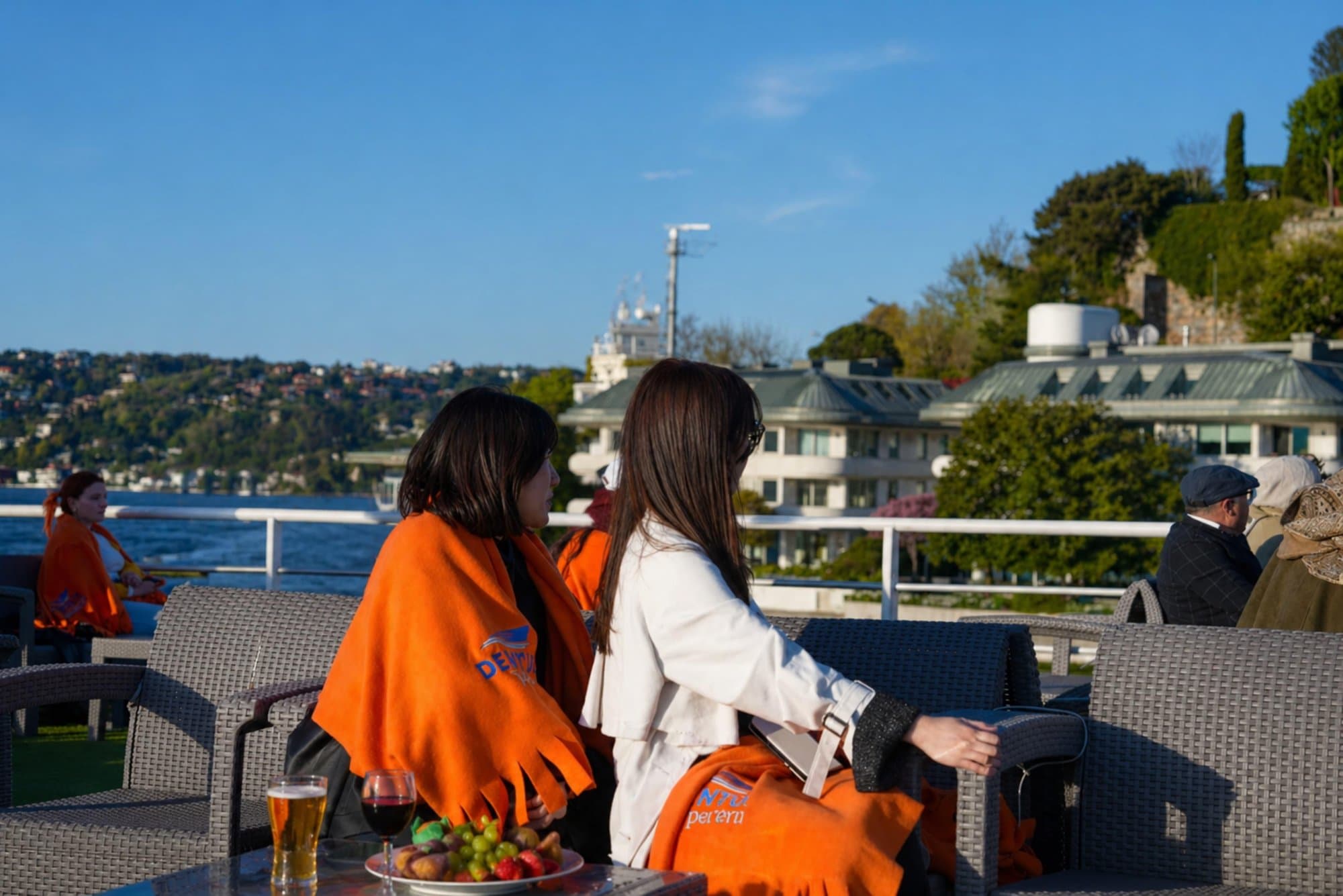 Guests relaxing on the open deck wrapped in blankets with drinks and fruit as the boat passes waterfront mansions