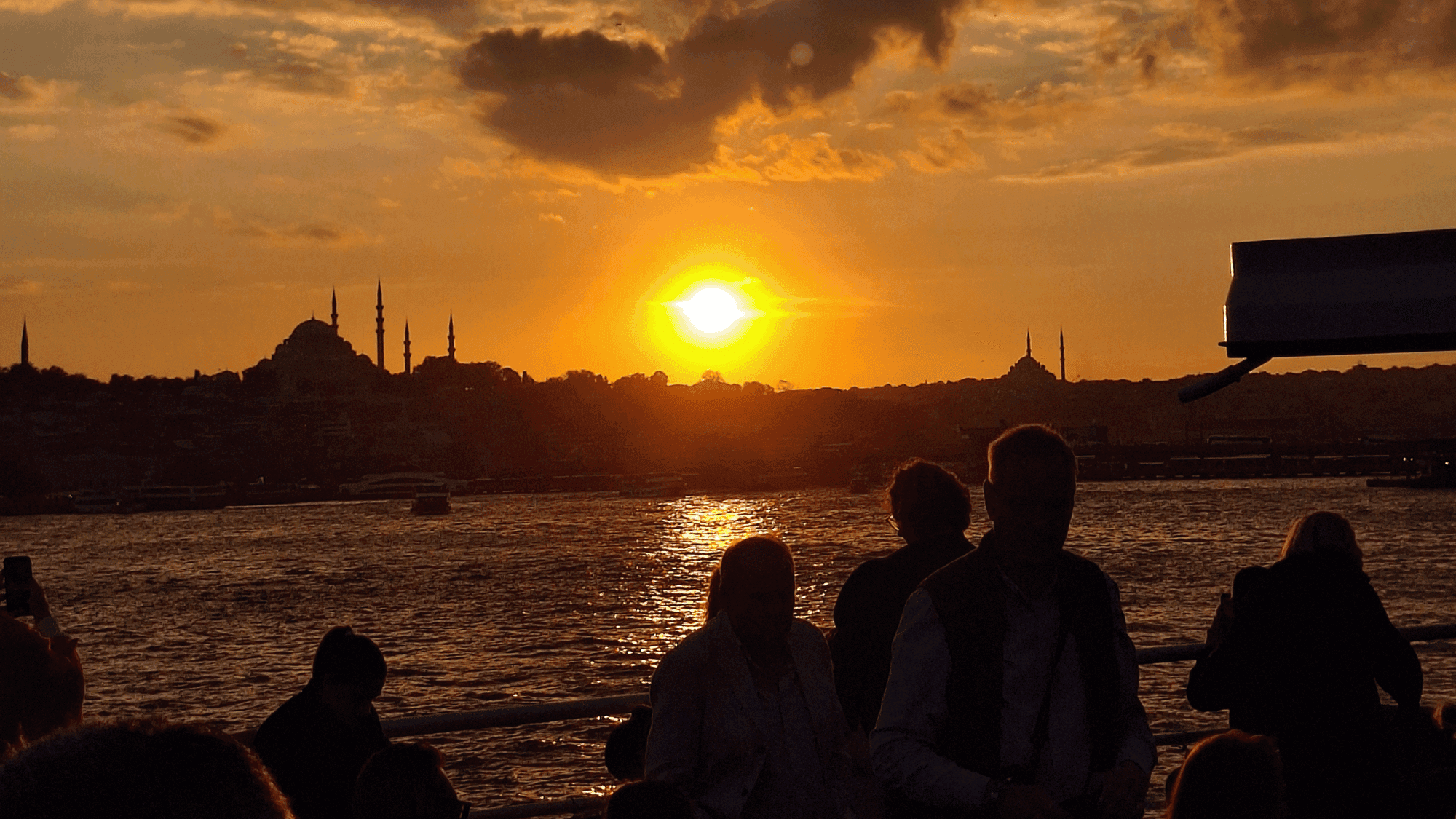 The sun setting behind the Süleymaniye Mosque silhouette as passengers watch from the boat deck
