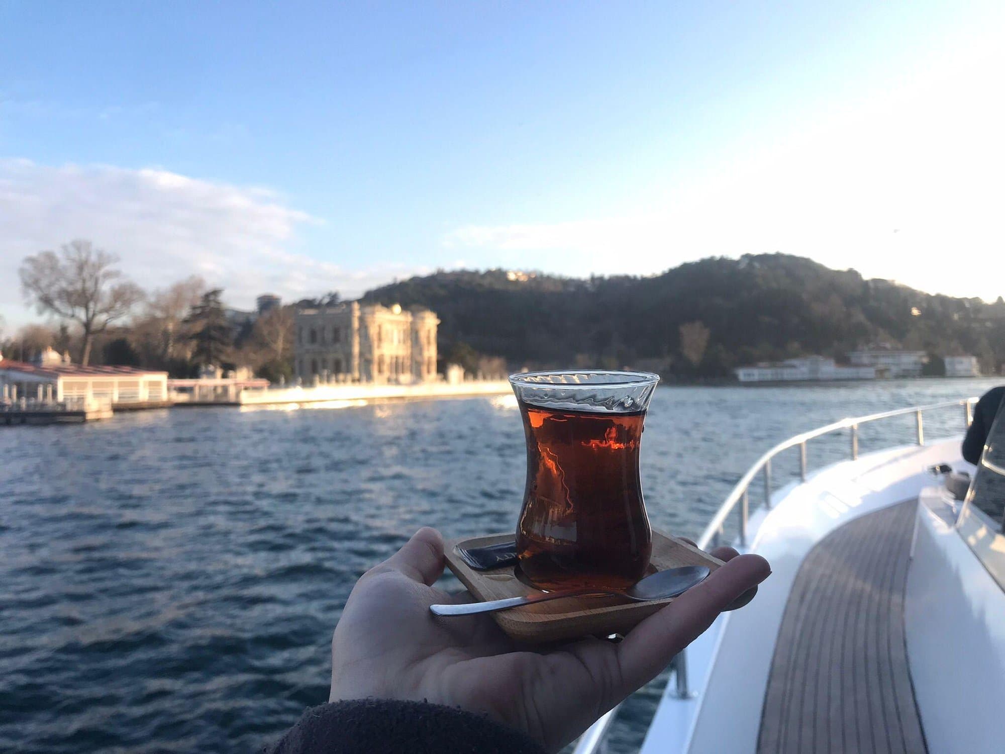 A glass of Turkish tea held up on deck with a waterside palace on the Bosphorus behind