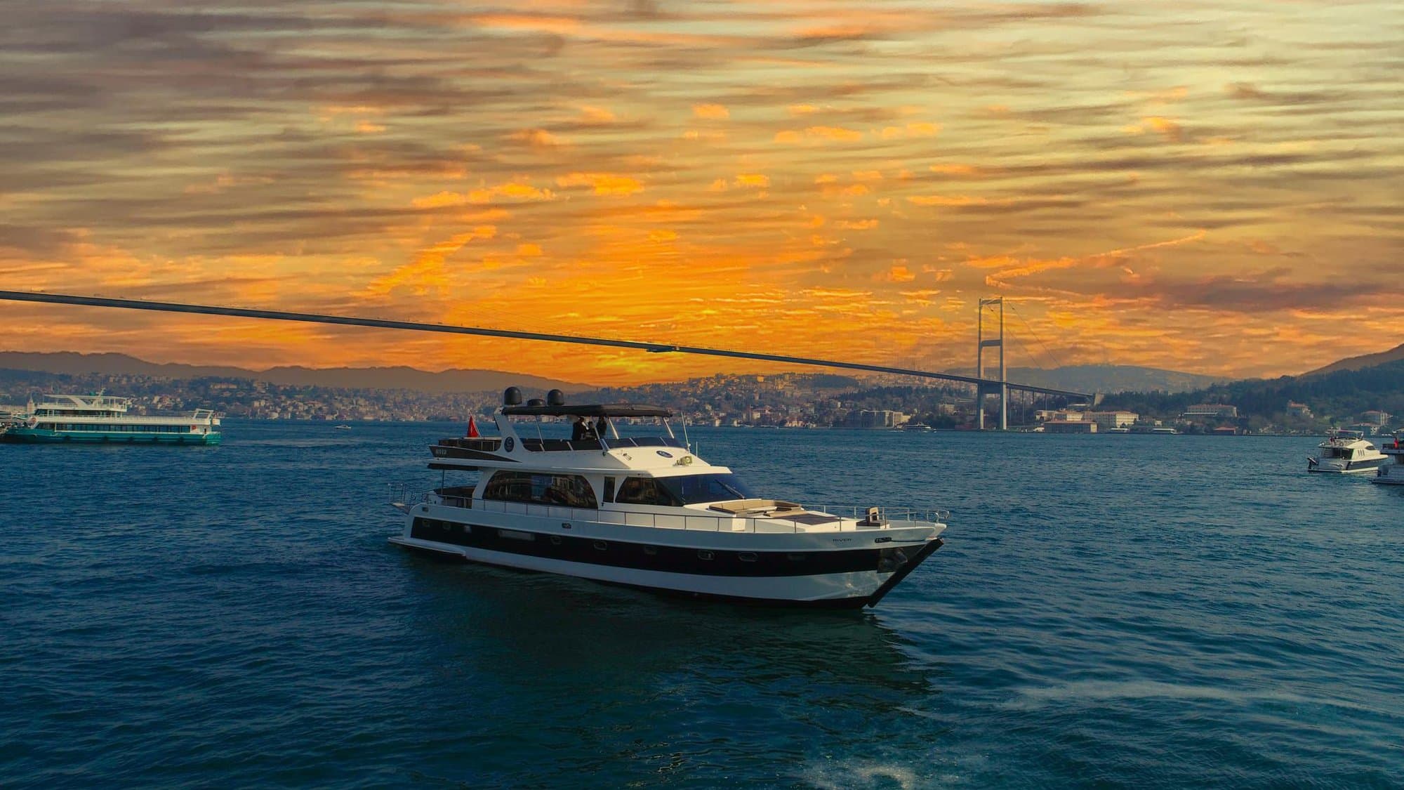A yacht cruising the Bosphorus past the Bosphorus Bridge under a glowing orange sunset