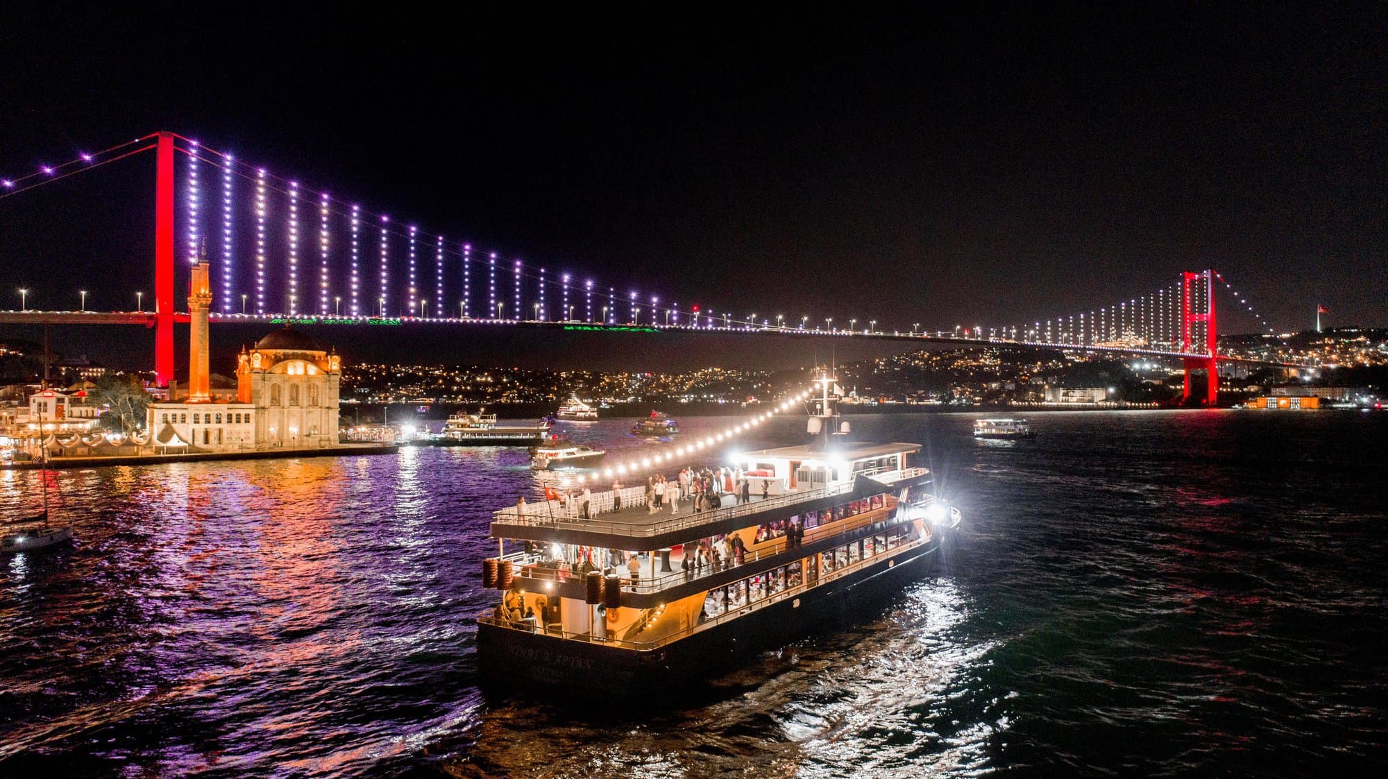 A dinner cruise boat lit up at night passing under the illuminated Bosphorus Bridge by the Ortaköy Mosque in Istanbul
