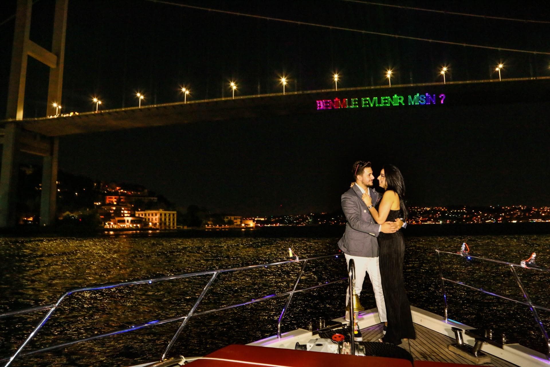 A couple on the yacht deck at night beneath the illuminated Bosphorus Bridge